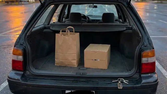 Open car trunk in a parking lot at dusk holding a closed paper bag and a sealed case of drinks