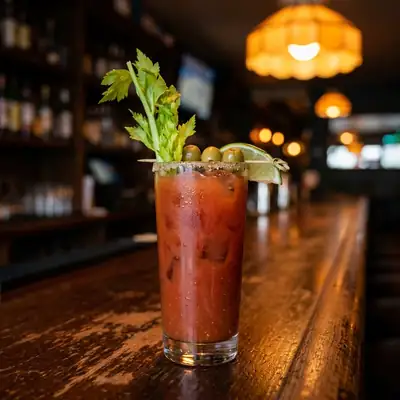 Caesar cocktail in a tall glass with a celery salt rim, celery stalk, lime wedge and olives on a wooden bar top